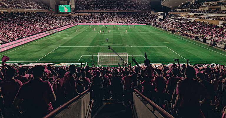 Blick von der Fantribüne auf ein Fußballspiel in einem vollbesetzten Stadion bei Nacht, Fans in Pink im Vordergrund.