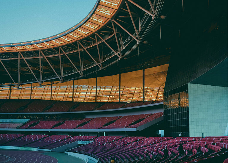 Interior of a large modern sports stadium at dusk with empty crimson seats, a sweeping illuminated roof canopy and a running track — evoking the scale of the global sports economy.